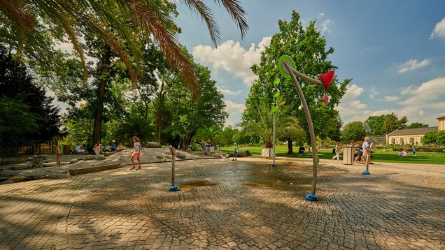 Kinder spielen auf dem Wasserspielplatz neben dem Haus Leonhardsbrunn. Palmen und andere Bäume umgeben den Spielplatz.