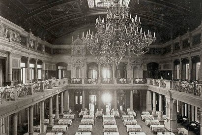 Eine geschichtliche Abbildung des Gesellschaftshauses, das sich am Palmenhaus anschließt. In der Halle stehen kleine Tische mit Stühlen und von der Decke hängt ein prunkvoller Kronleuchter.