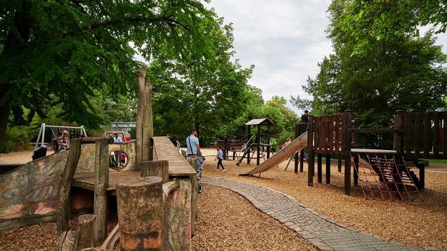 Der Spielplatz mit einigen Klettergerüsten aus Holz, umgeben von Bäumen.