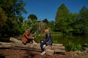 Auf einem Baumstamm sitzen zwei Frauen. Sie wenden der Kamera ihren Rücken zu. Der Baumstamm steht vor einem Weiher. Dieser besitzt einen Wasserfall. Der Fernsehturm zeigt sich im Hintergrund.