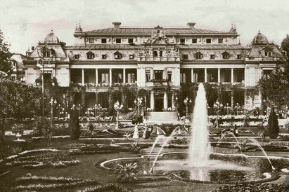 Eine alte Abbildung des Palmenhauses. Die Abbildung ist in schwarz-weiß gehalten. Im Hintergrund befindet sich das Palmenhaus. Im Vordergrund ist ein Garten, in dem sich ein großer Springbrunnen befindet. Zwischen dem Palmenhaus und dem Garten befindet sich ein weg, der mit vielen Laternen versehen ist.