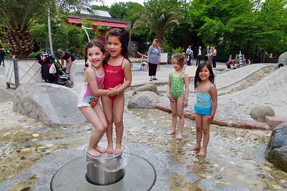 Kinder spielen fröhlich am Wasserspielplatz in Badekleidung. Im Hintergrund befinden sich Eltern und Familien.