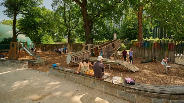 ein Spielplatz mit vielen Holzgerüsten ist von einigen Bäumen umgeben. Ein paar Kinder spielen auf dem Spielplatz. Einige Eltern schauen zu und sind auch auf dem Spielplatz.