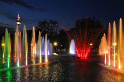Ein Brunnen mit Wasserfontänen ist mit bunten Lichtern beleuchtet. Die Sonne ist schon untergegangen und der Himmel ist dunkel.