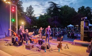 Auf der Bühne des Musikpavillons steht eine Gruppe von Musiker:innen. Darunter befinden sich ein Sänger, Gitarrenspieler und Schlagzeuger. Ein großes Publikum sitzt ihnen gegenüber. Um die Zuschauer herum stehen viele Bäume.